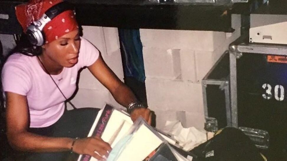 Young Honey Dijon sits beside DJ equipment in New York, sorting vinyl records while wearing headphones and a red bandana.