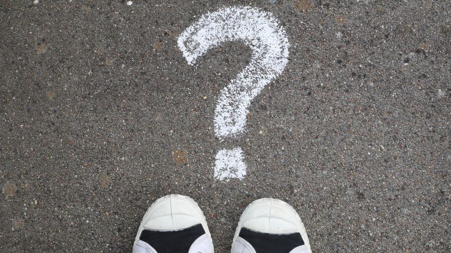 Top-down view of shoes facing a large white question mark drawn on a paved surface outdoors.