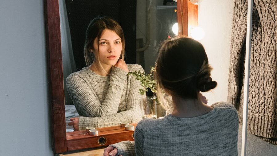 Young woman seated at a dressing table, gazing into a mirror beside a small plant and decorative lights.