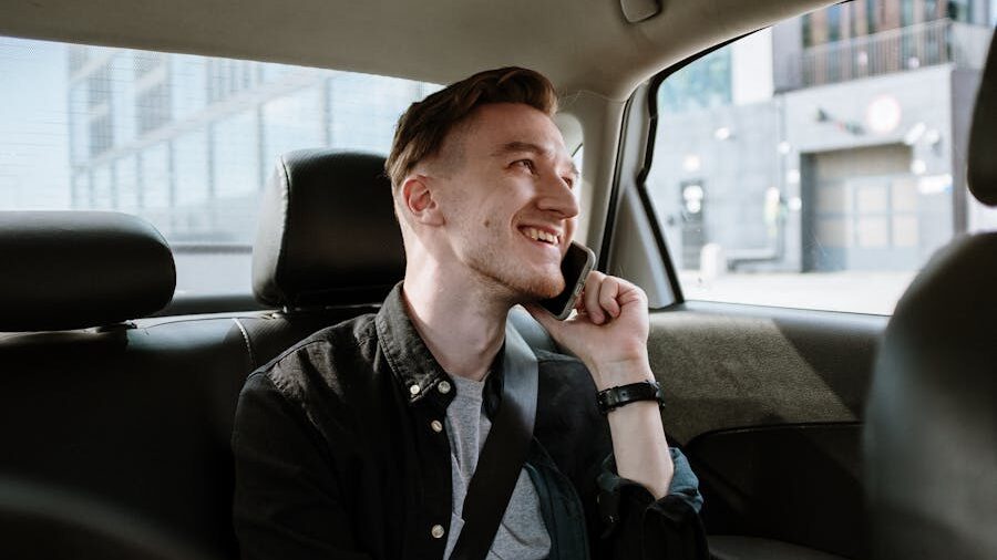 Young man seated in the back seat of a car, smiling while holding a smartphone to his ear and looking outside.