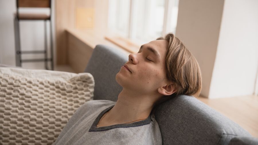 Individual leaning back on a couch with eyes closed, appearing relaxed near a window in a home setting.