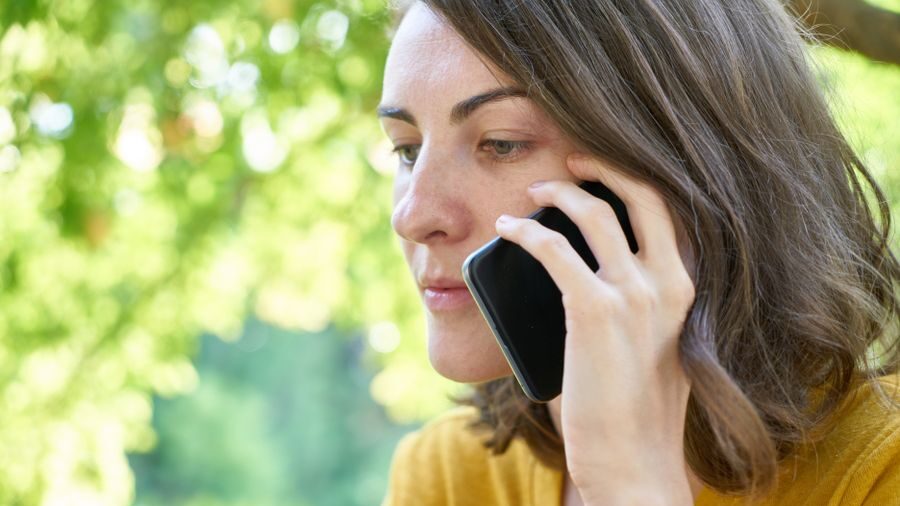 Outdoor portrait of a woman in a yellow top holding a smartphone to her ear while listening during a call.