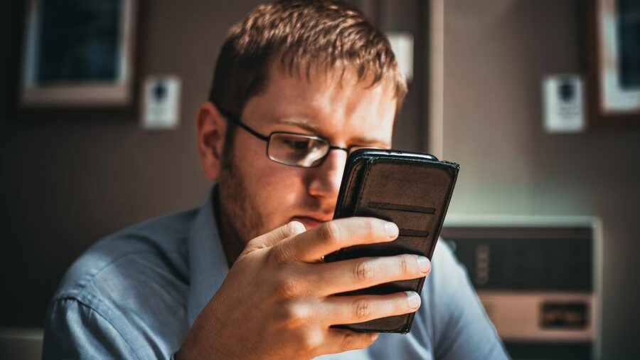 Indoor portrait of a man using a smartphone, wearing glasses and focusing on content displayed on the screen.