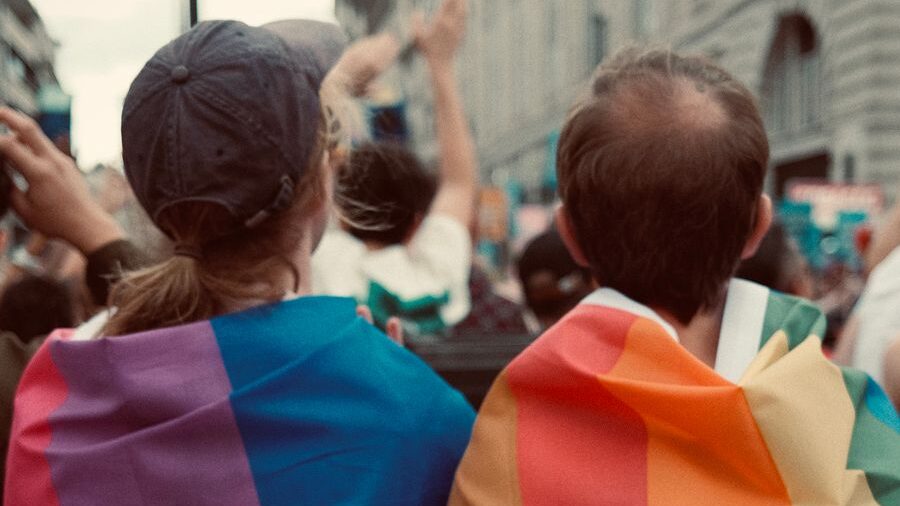 Two people viewed from behind wearing rainbow flags over their shoulders while attending a crowded pride event.