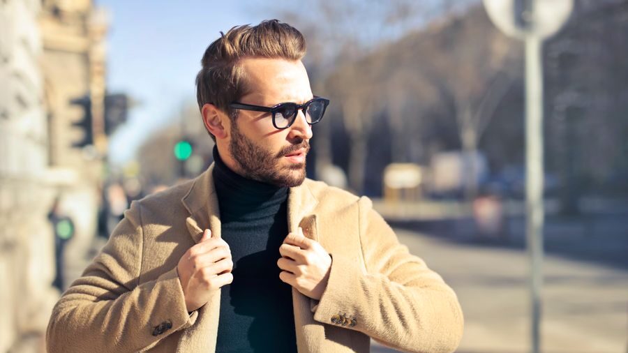 Bearded man in sunglasses and a beige overcoat standing outdoors on a sunlit street with blurred buildings behind him.