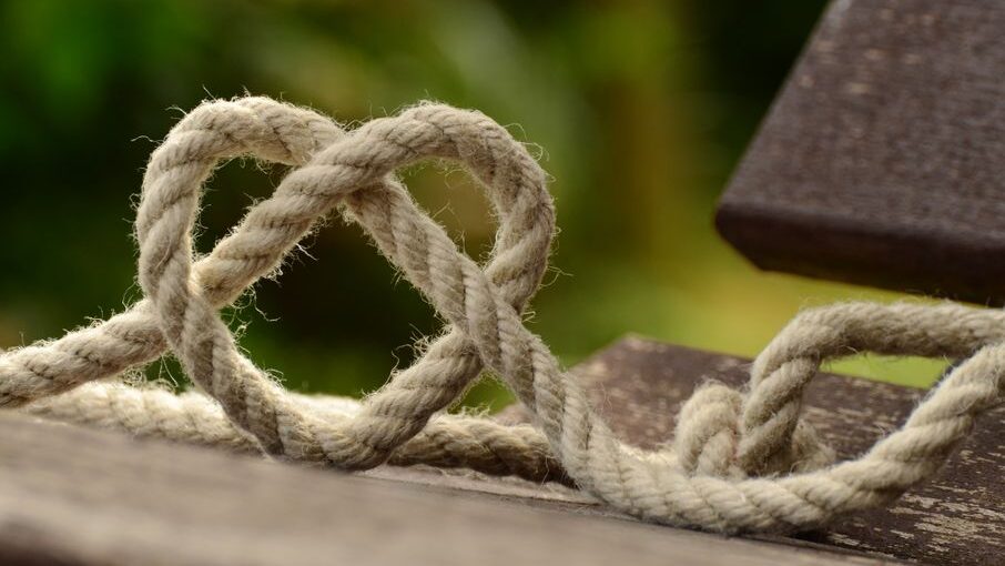 Close-up of a rope twisted into a heart shape on a wooden bench, with a blurred green background outdoors.