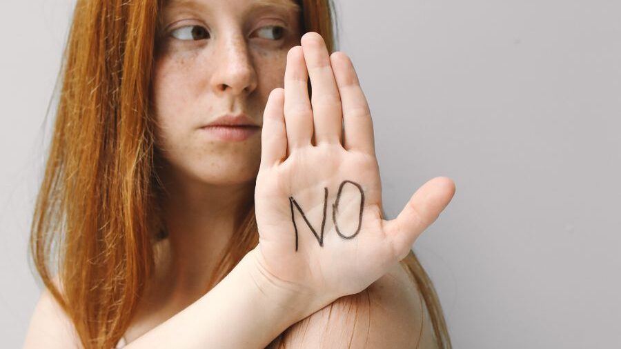 Red-haired woman displaying a stop gesture with the word “NO” on her hand against a plain light background.
