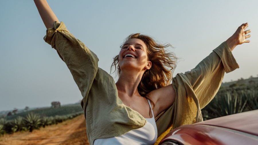 Woman wearing a light jacket smiles with outstretched arms next to a car, surrounded by an open landscape.