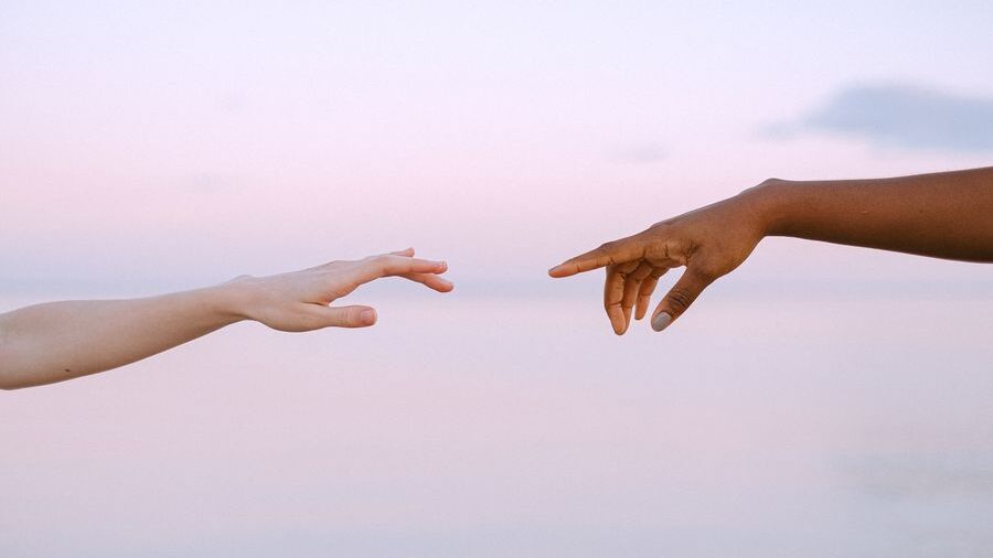 Two outstretched hands reaching toward each other against a soft pastel sky, with water and horizon in the background.