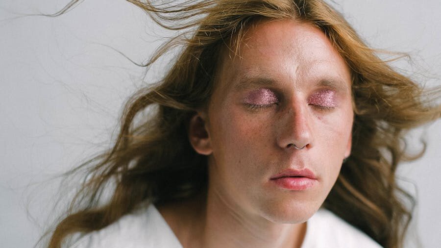 Close-up portrait of a person with long flowing hair, closed eyes, glitter eye makeup, and a nose ring against a light background.