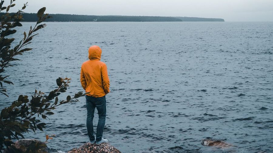 Individual standing near a lake in a yellow jacket and jeans, facing the water with a distant tree-lined shore ahead.