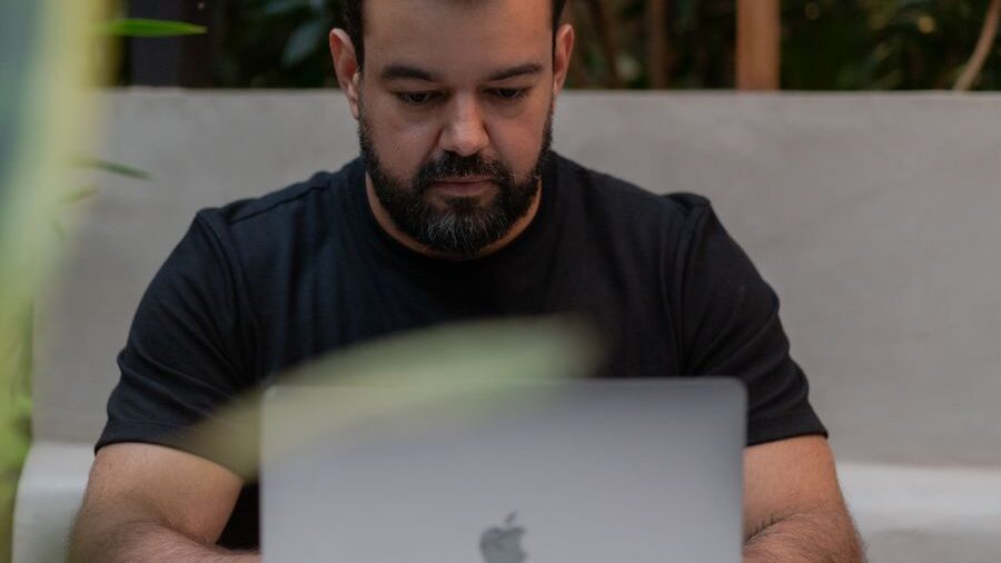 Man seated at a table using a laptop computer, with blurred plants visible in the foreground and background.