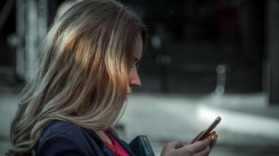 Side view of a woman with long blonde hair looking at a smartphone while standing outdoors in an urban setting.