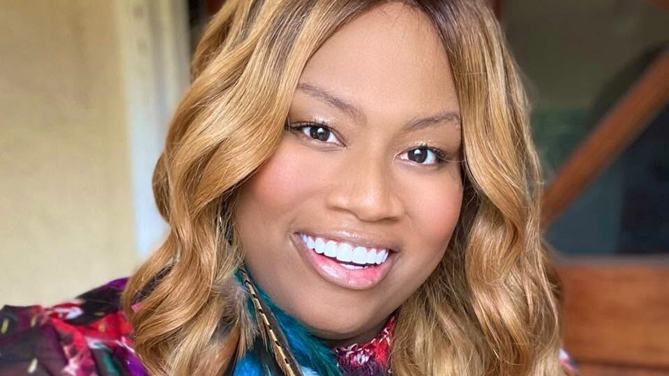 Close-up portrait of Ashlee Marie Preston wearing a floral outfit, statement feather earrings, and long blonde waves while smiling at the camera.