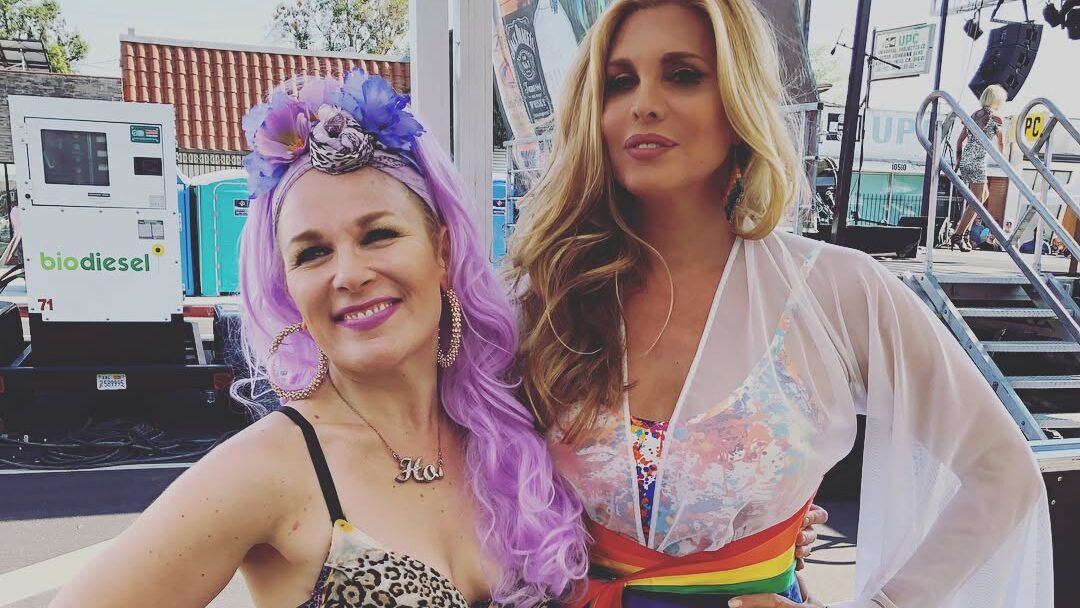 Candis Cayne posing with actress Wendy Benson-Landes at Valley Pride, both smiling outdoors in colorful outfits during the celebration.