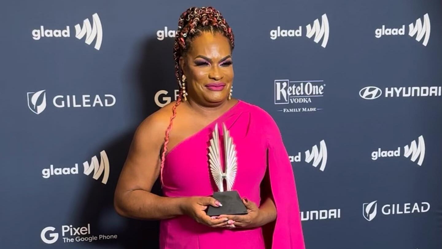 Imara Jones poses in a bright pink one-shoulder gown while holding an award at the GLAAD Media Awards in front of the event sponsor backdrop.