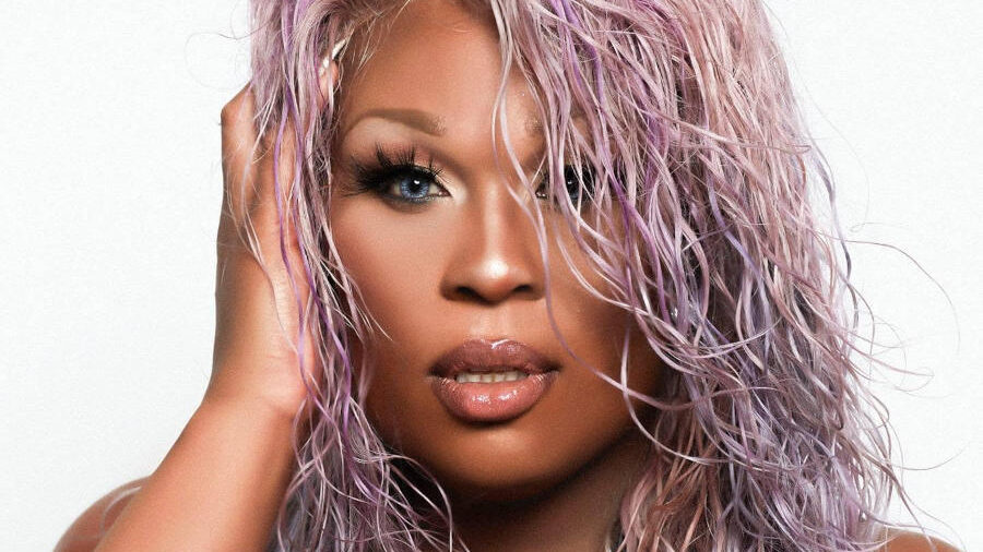 Portrait of Peppermint with pastel lavender wet-look hair, dramatic eye makeup, and a black outfit as she poses against a white background.