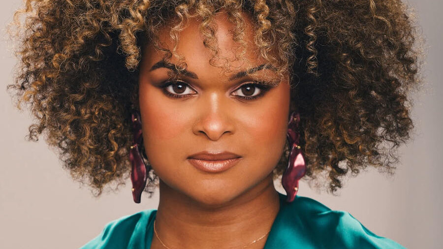 Portrait of Raquel Willis wearing a teal satin blouse, statement earrings, and a short curly hairstyle while looking directly at the camera.