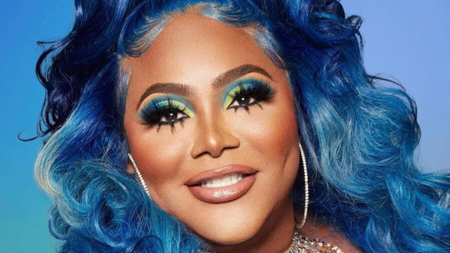 TS Madison wearing a vibrant blue outfit and matching blue wavy hair, accessorized with large hoop earrings and a sparkling 