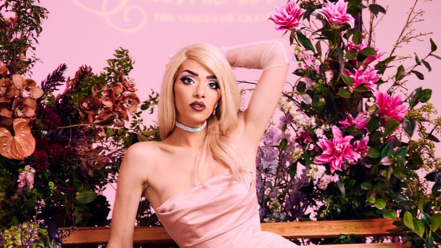 Arisce Wanzer wearing a pink satin gown while posing on a wooden bench surrounded by pink flowers and decorative plants.