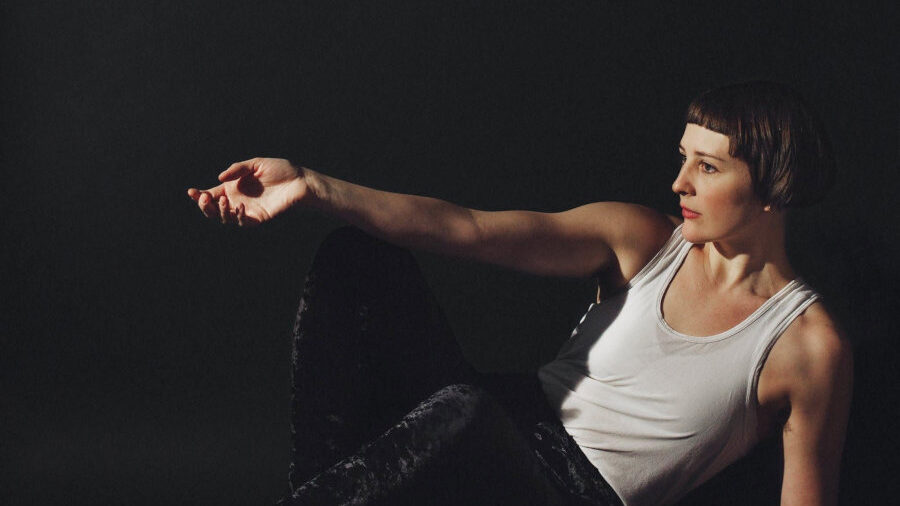 Kade Pyle seated against a dark background, wearing a white tank top and black pants with one arm extended.