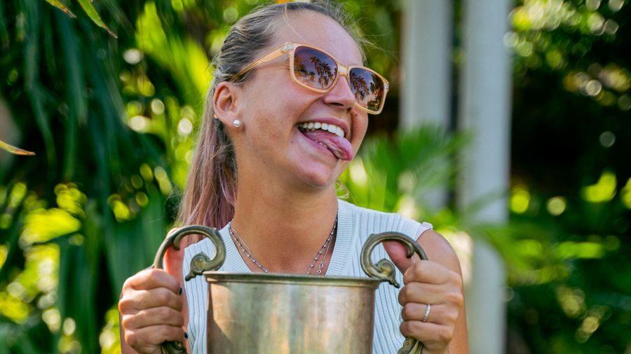 Woman wearing sunglasses smiling and holding a large silver trophy with both hands in a green outdoor setting