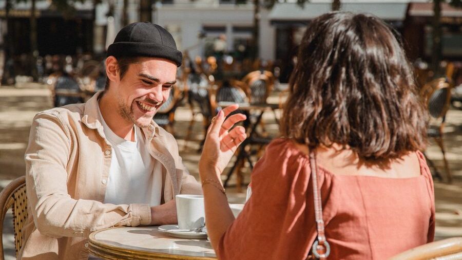 Two people smiling and talking over coffee while sitting together at an outdoor café terrace on a sunny day.