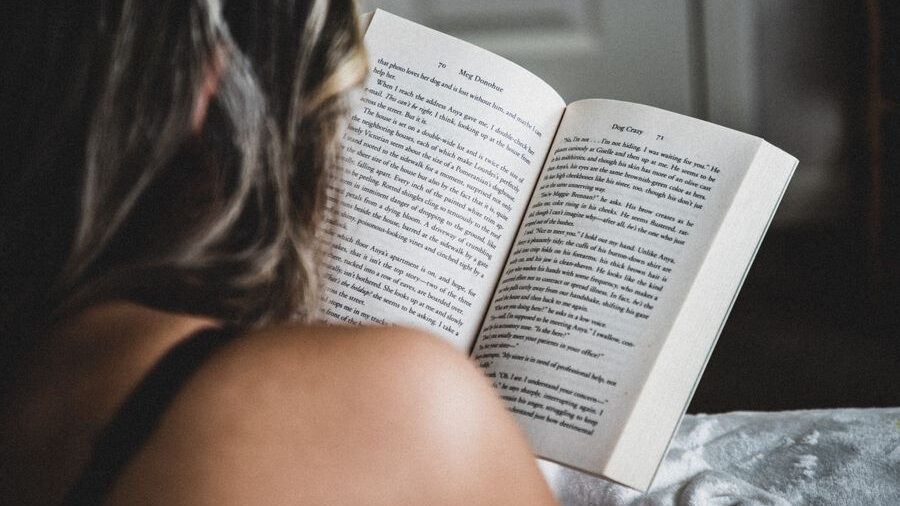 Person with long dark hair reading an open book while sitting on a bed, photographed from behind in soft natural light