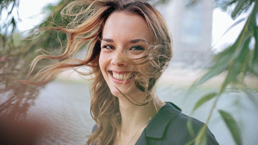 Close-up portrait of a smiling woman with long brown hair, with loose strands of hair falling across her face