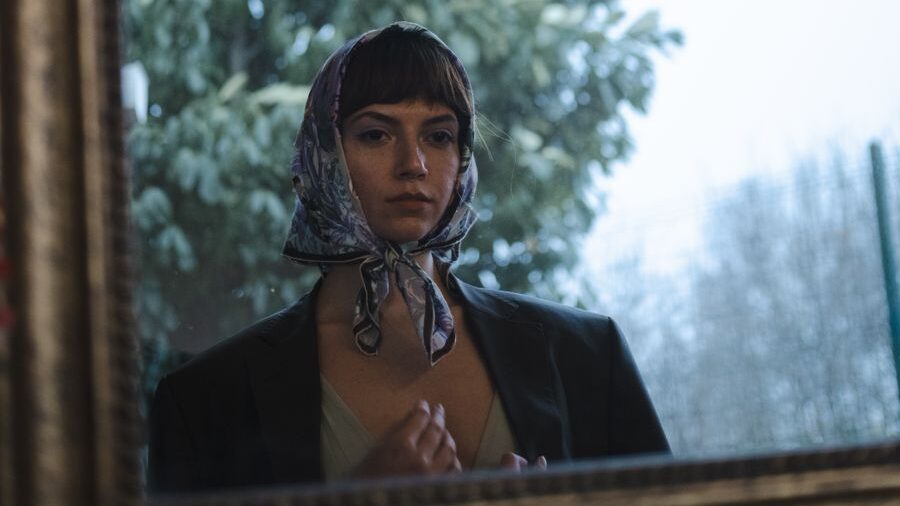 A woman wearing a patterned headscarf and dark jacket looks at her reflection in a mirror beside a large window.
