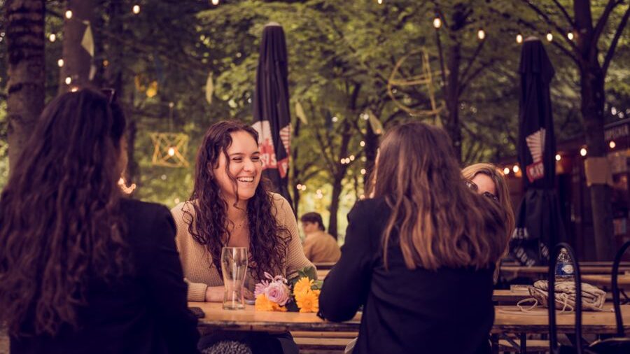 A group of friends sitting around an outdoor café table, smiling and talking together beneath trees and string lights.