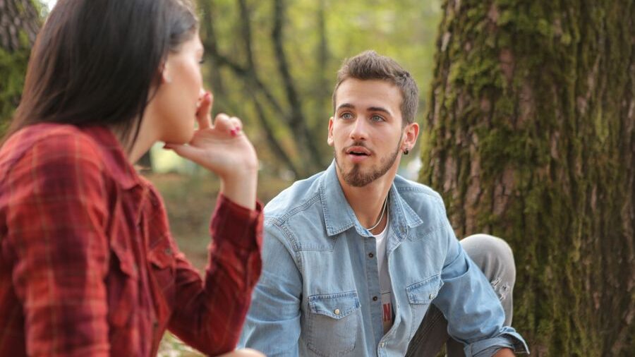 A man and woman sitting outdoors beside a large tree and facing each other while having a conversation in a wooded area.