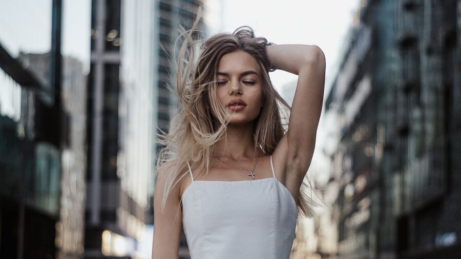 Young woman in a short white dress posing on a city street with tall modern buildings and traffic in the background