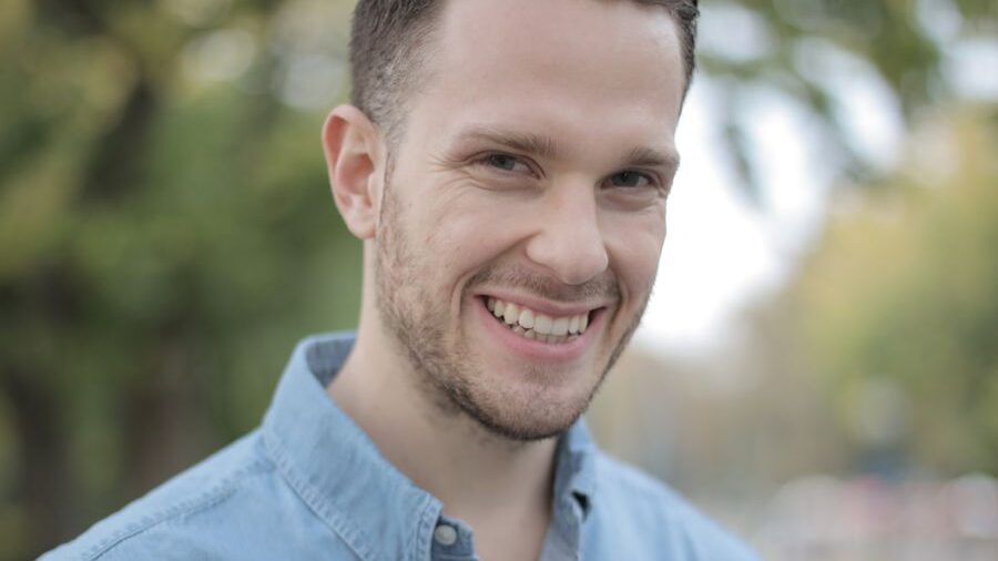 Close-up portrait of a smiling man with short brown hair and light facial hair, wearing a blue shirt outdoors.