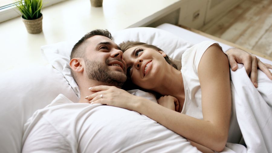 Smiling couple lying together in bed, with the woman resting her head on the man’s shoulder and embracing him.