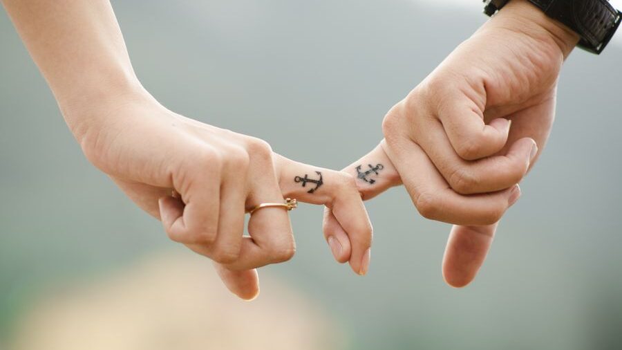 Two hands making a pinky promise, showing matching anchor tattoos on their fingers, with a blurred background.