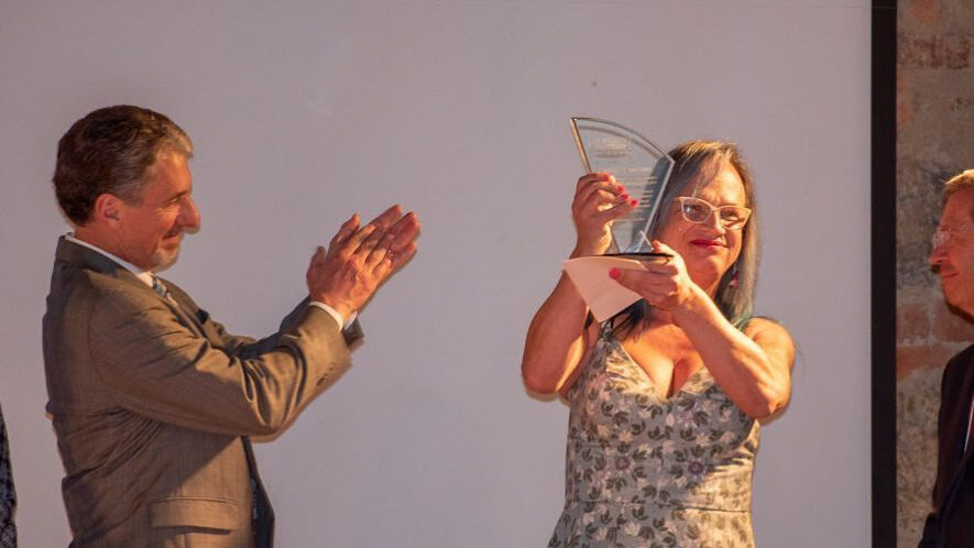 Brigitte Baptiste, con pelo largo y gafas, sosteniendo un premio mientras sonríe. Un hombre de traje la aplaude en el escenario