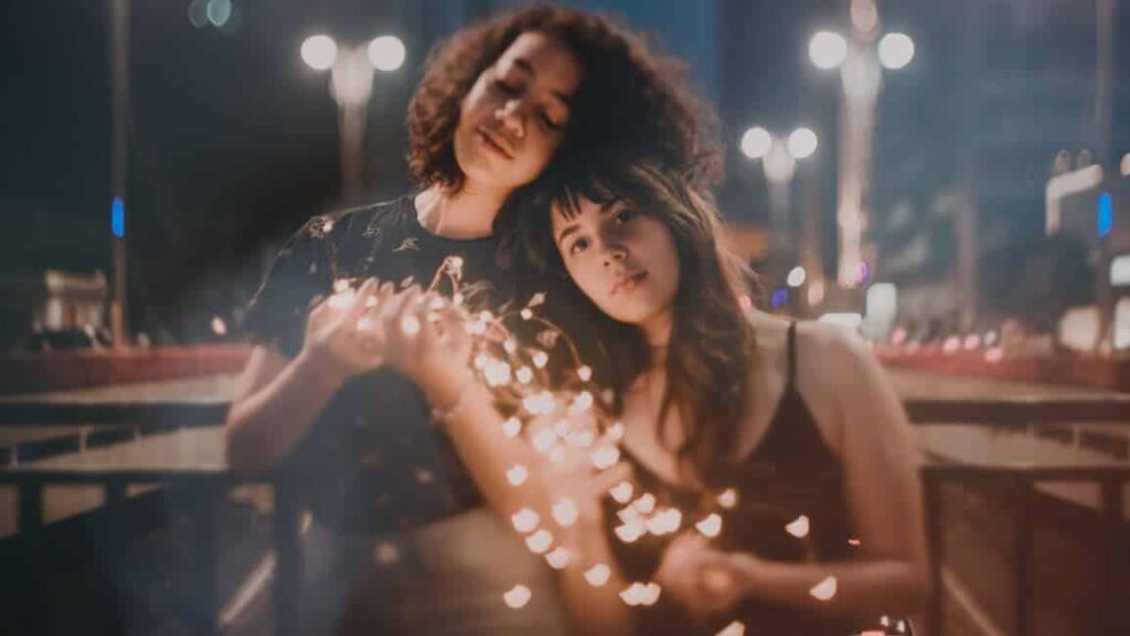 Dos mujeres jóvenes sostienen una tira de luces, apoyadas la una en la otra en una calle de la ciudad por la noche, rodeadas de cálidos corazones borrosos.