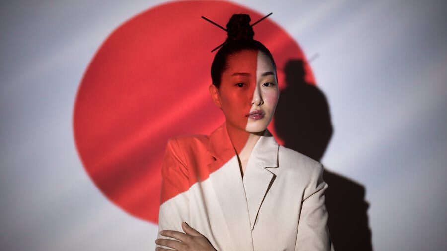 Una mujer con un peinado minimalista y palillos en su cabello, de pie frente a una bandera japonesa proyectada.