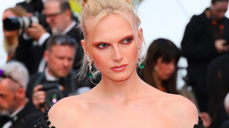 Alex Consani en una alfombra roja luciendo un vestido negro con los hombros descubiertos y adornado con cuentas brillantes, con el pelo recogido con elegancia y aretes verdes.