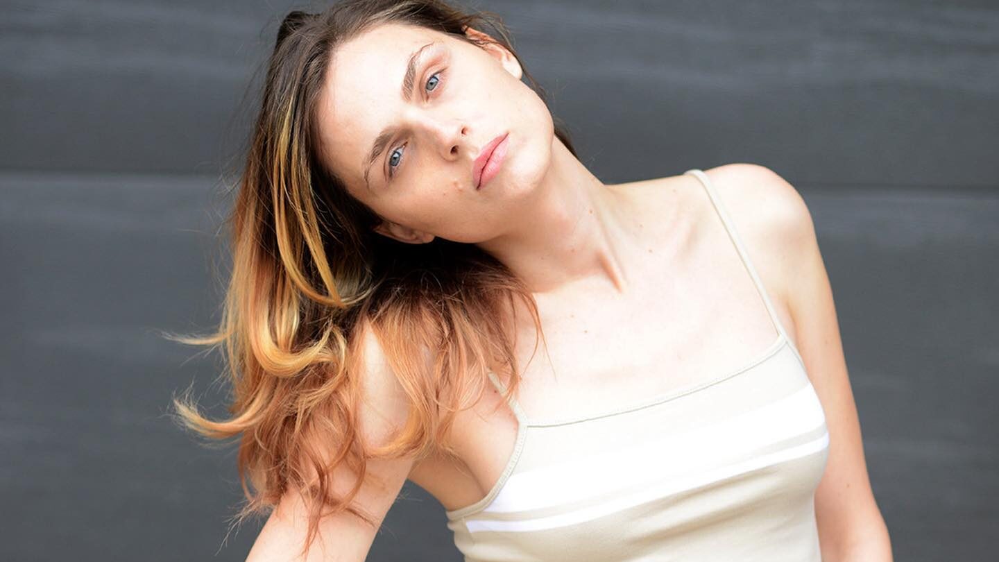 Andreja Pejić frente a un fondo gris, con una camiseta corta de tirantes blanca y vaqueros blancos de cintura alta, con la cabeza inclinada y el cabello sobre un hombro.