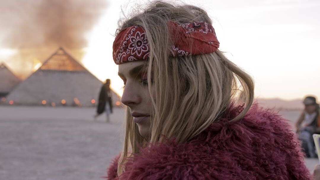 Andreja Pejić en el desierto de Burning Man, en 2016, vestida con un pañuelo rojo y un abrigo de piel sintética, con el fuego brillando detrás de ella.