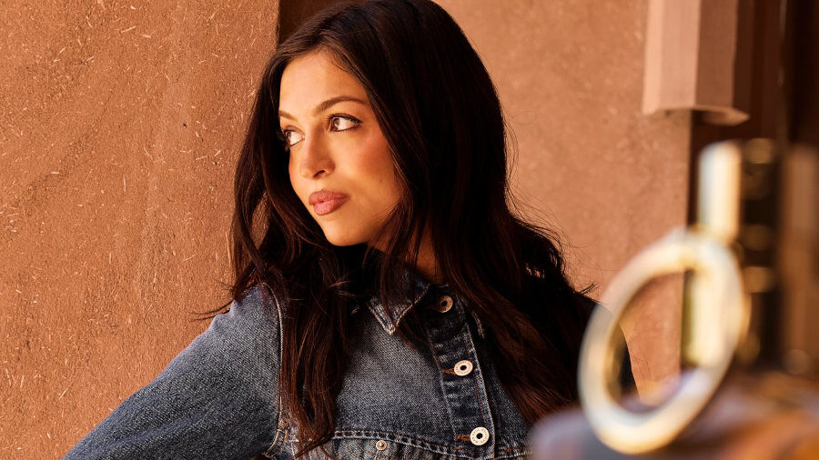 Josie Totah, con una chaqueta de mezclilla y jeans de Jean Paul Gaultier, posa apoyada contra una pared beige con textura.