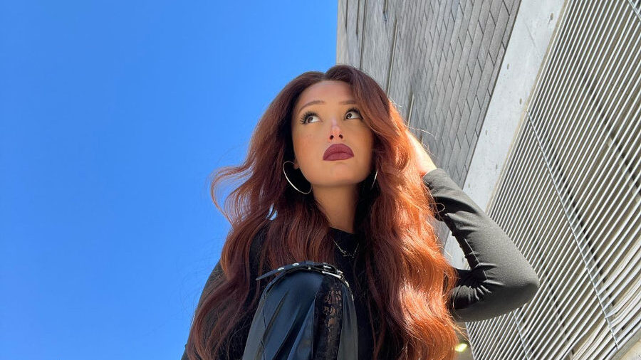 Zoey Luna posa al aire libre con pantalones de cuero y cabello colorado con ondas, sentada en un sentada sobre un contenedor bajo el sol.