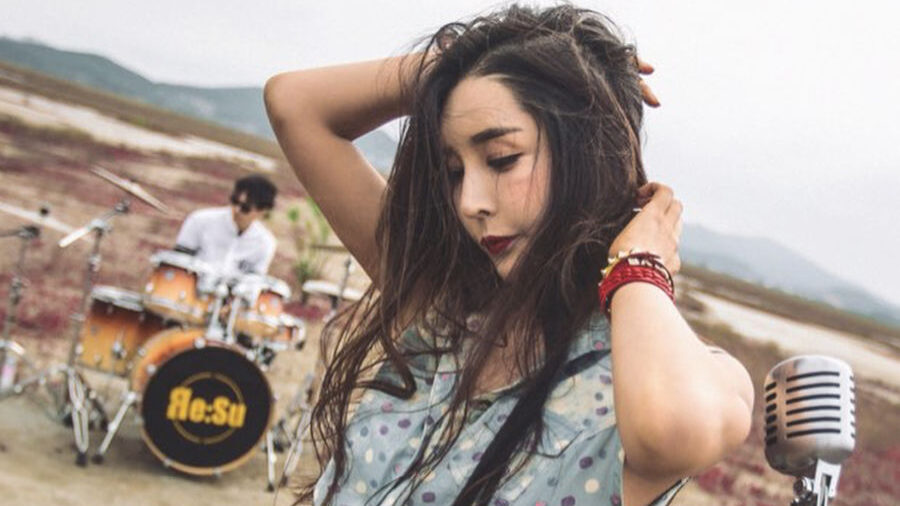 Harisu posa con un micrófono vintage frente a un baterista, en un amplio paisaje al aire libre con hierba roja.