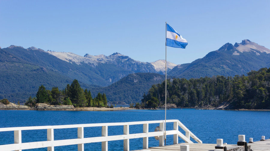 Bandera argentina en un muelle con vistas a un lago azul y montañas boscosas en la Patagonia bajo un cielo celeste.
