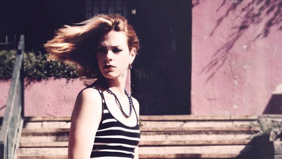 Una joven Daniela Vega con una camiseta a rayas sentada en unas escaleras con azulejos mientras el viento le sopla el cabello.