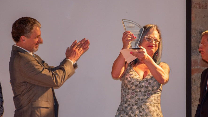 Brigitte Baptiste brandissant une plaque de récompense en souriant. Un homme en costume l'applaudit sur scène.