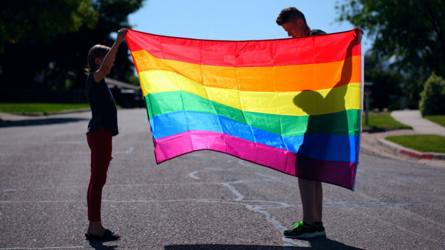 Deux personnes brandissent fièrement un drapeau arc-en-ciel, projetant des ombres sur le sol.