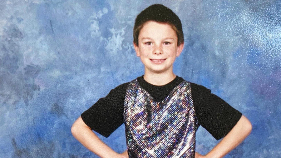 Dylan Mulvaney enfant en gilet brillant et tenue noire, prenant une pose confiante avec les mains sur les hanches, dans un studio.